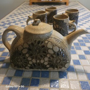 May include: A ceramic teapot with a matching lid and four mugs. The teapot and mugs are beige with a textured floral pattern. The set is on a blue and white checkered surface.
