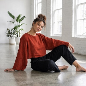 Puede incluir: Una blusa de manga larga de color &oacute;xido con mangas anchas y pantalones negros. La persona est&aacute; sentada en un suelo de hormig&oacute;n frente a una pared de ladrillos blancos con grandes ventanas y plantas en macetas.