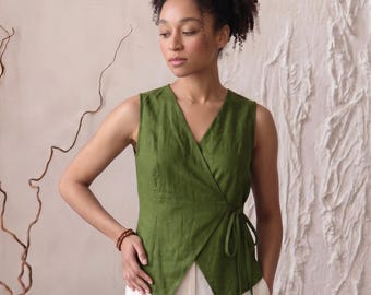 May include: A sleeveless olive green wrap top featuring a V-neckline and a side tie closure, paired with loose-fitting white trousers. The top is constructed with a fitted bodice and a pointed hemline that overlaps at the front waist.