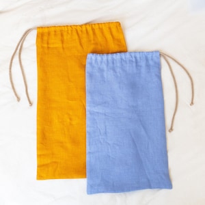 May include: Two rectangular drawstring bags. One is a vibrant yellow-orange, the other a soft blue. Both are made of linen-like fabric and have a natural-colored drawstring closure. The bags are displayed on a white surface.