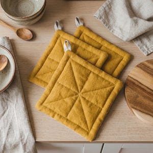 May include: Three square, mustard-yellow quilted pot holders with white hanging loops. The pot holders are arranged on a light-colored wooden surface, alongside a wooden spoon, bowls, and a cutting board. A linen napkin is also present.