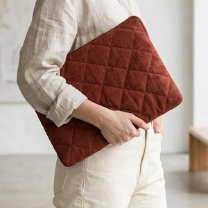Puede incluir: Una funda acolchada para portátil, de color óxido, con un patrón de diamantes. La funda la sostiene una persona que lleva una camisa beige abotonada y pantalones color crema. El fondo incluye una ventana, un banco y una pintura.