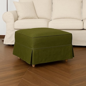 Op de afbeelding: Een olijfgroene poef met een geplooide rok staat voor een crèmekleurige bank. De poef heeft een vierkante vorm en rust op korte, lichtgekleurde houten poten. Links staat een vloerlamp met een beige kap.