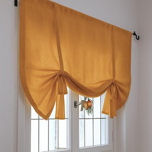 Op de afbeelding: Mosterdkleurig Romeins rolgordijn, vastgebonden met bijpassende stoffen banden. Het gordijn hangt boven een wit raam met een rasterpatroon. Het raam staat in een witte muur.