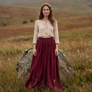 May include: A long-sleeved, light beige button-down shirt paired with a floor-length, deep burgundy skirt. The shirt features a simple collarless neckline and a buttoned front, while the skirt has a gathered, full-length silhouette.