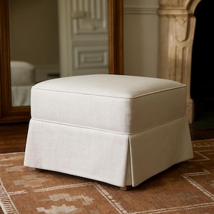 May include: A rectangular, off-white upholstered ottoman with a skirted base. The ottoman has a tailored appearance with a piped edge and sits on short, square feet. The neutral color and classic design suggest a living room or bedroom furniture piece.