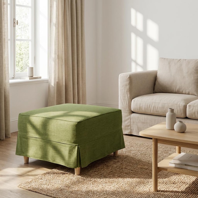 May include: A square, olive green ottoman with a skirted base and light wooden legs. The ottoman is in a living room setting, with a beige sofa, wooden coffee table, and natural fiber rug.