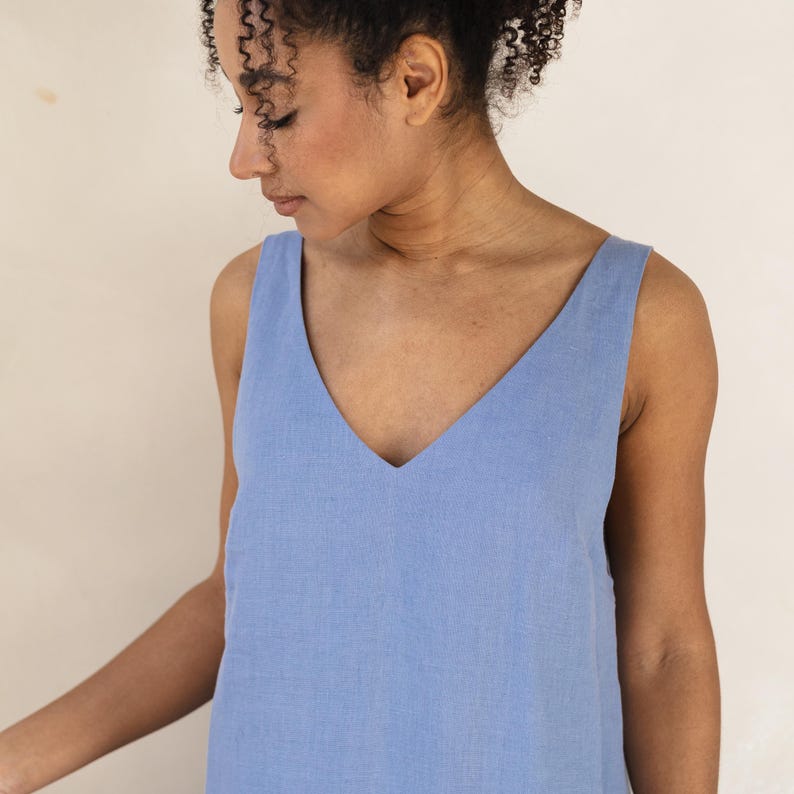 May include: A light blue sleeveless dress with a deep V-neck. The dress is made of a textured fabric and has a simple, elegant design. The model is wearing the dress.