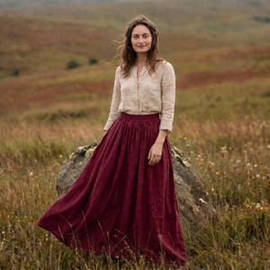 May include: A long-sleeved beige button-down shirt paired with a floor-length, deep burgundy skirt. The skirt features a gathered, high-waisted design that flows into a full, voluminous silhouette, reaching down to the ankles in a solid colour.
