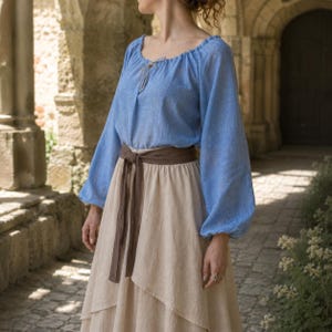 May include: A light blue long-sleeved blouse with a gathered neckline and a thin tie closure, paired with a beige layered skirt featuring a wide dark brown fabric belt tied at the waist. The blouse has loose sleeves and a textured fabric finish.