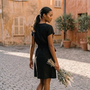 Puede incluir: Un vestido cruzado negro con mangas cortas y cintur&oacute;n a juego. El vestido llega a la rodilla y lo lleva una persona que sostiene un ramo de flores secas. El fondo presenta una calle adoquinada y edificios con contraventanas.
