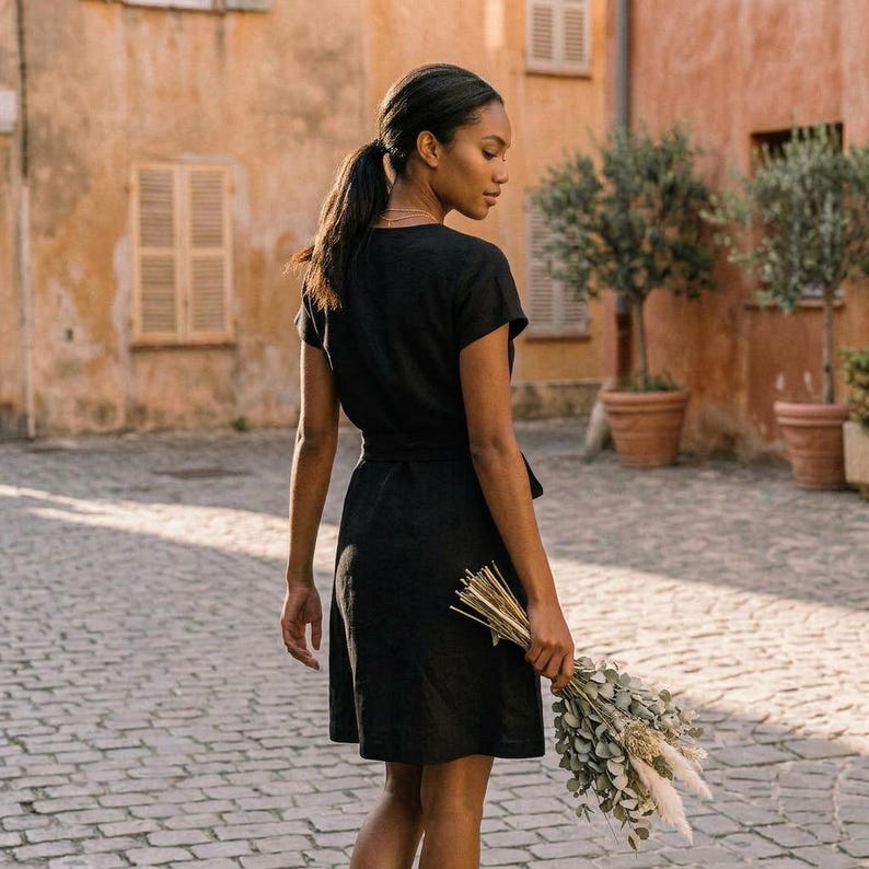 Puede incluir: Un vestido cruzado negro con mangas cortas y cintur&oacute;n a juego. El vestido llega a la rodilla y lo lleva una persona que sostiene un ramo de flores secas. El fondo presenta una calle adoquinada y edificios con contraventanas.