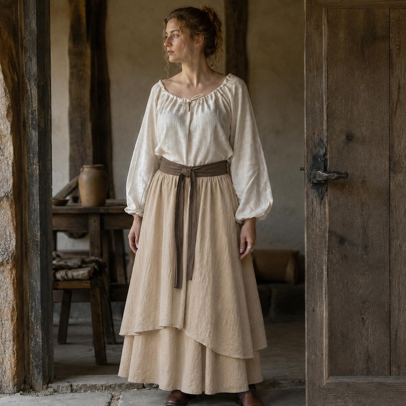 May include: A long-sleeved off-white blouse with a gathered neckline and tie closure, paired with a layered beige maxi skirt and a dark brown fabric waist sash. The ensemble features a textured, woven appearance with a loose, flowing silhouette.