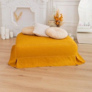 Op de afbeelding: Een mosterdgele ottoman met een geplooide rok, bekroond met drie kussens in crème- en beigetinten. De ottoman is rechthoekig en staat op een lichte houten vloer. Decoratieve elementen zijn kaarsen en een spiegel.