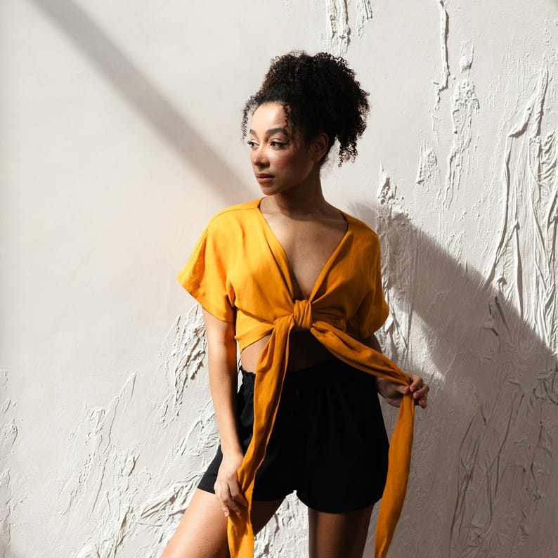 May include: A mustard yellow tie-front top with short sleeves and a V-neck. The top is worn with black shorts. The model is positioned in front of a textured white wall, with shadows from sunlight.