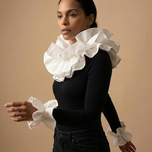Op de afbeelding: Een zwart topje met lange mouwen met een grote witte gerimpelde kraag en bijpassende gerimpelde manchetten. Het model draagt een zwarte jeans. De achtergrond is effen beige. De top is een uniek fashion statement.