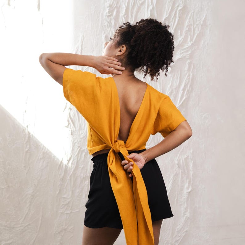 May include: Mustard yellow wrap top with a deep V-neck and short sleeves. The back is open, with long ties that wrap around the waist and tie in a knot. Worn with black shorts.