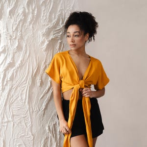 May include: Mustard yellow wrap top with short sleeves and a deep V-neck. The top is tied at the waist with long, flowing ties. Paired with black shorts. The top is made of a soft, flowing fabric.