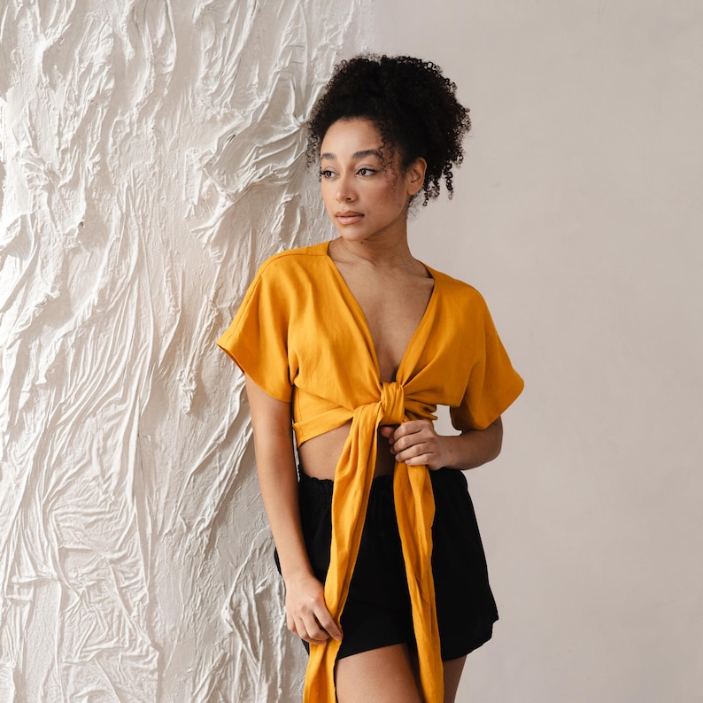 May include: Mustard yellow wrap top with short sleeves and a deep V-neck. The top is tied at the waist with long, flowing ties. Paired with black shorts. The top is made of a soft, flowing fabric.
