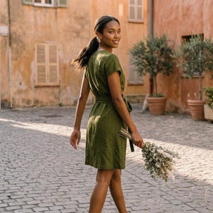 Puede incluir: Una mujer con un vestido verde oliva de manga corta y cintur&oacute;n a juego. Sostiene un ramo de flores secas y camina por una calle adoquinada. El fondo muestra edificios con contraventanas y &aacute;rboles en macetas.