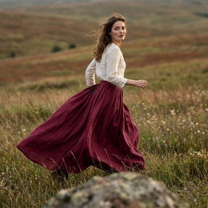 May include: A long-sleeved cream-coloured blouse paired with a voluminous, floor-length deep burgundy skirt. The skirt features a gathered waistline and a full, flowing silhouette that extends to the ground, crafted from a textured fabric.
