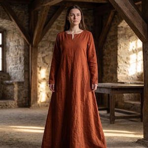 May include: A long, rust-colored dress with long sleeves and a V-neck. The dress is made of a flowing fabric and is worn by a person standing in a room with stone walls and wooden beams. The dress has a simple, medieval design.
