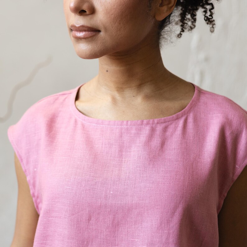 May include: A pink, short-sleeved top with a round neckline. The fabric appears to be linen or a similar woven material. The top is a solid color, with a subtle texture visible in the close-up shot.