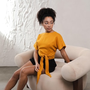 May include: Mustard yellow wrap top with short sleeves and a tie closure. The top is paired with black shorts. The model is seated in a beige curved armchair against a white textured wall.