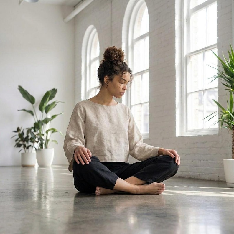 Puede incluir: Una persona con una blusa beige de manga larga y pantalones negros est&aacute; sentada con las piernas cruzadas en un suelo de hormig&oacute;n. La habitaci&oacute;n tiene grandes ventanas y plantas en macetas. La blusa tiene cuello redondo y los pantalones son holgados.