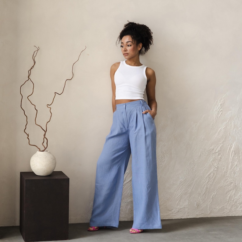 May include: A woman wearing a white tank top and light blue wide-leg pants. She is standing in front of a white wall with a vase of dried branches on a black pedestal.