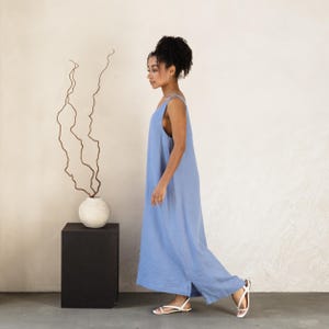May include: A light blue jumpsuit with wide legs and thin straps. The model is wearing white sandals. The jumpsuit is made of a lightweight fabric. A white vase with dried branches sits on a black pedestal.