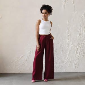 May include: A woman wearing a white tank top and wide-legged burgundy pants. The pants have a high waist and pleats.
