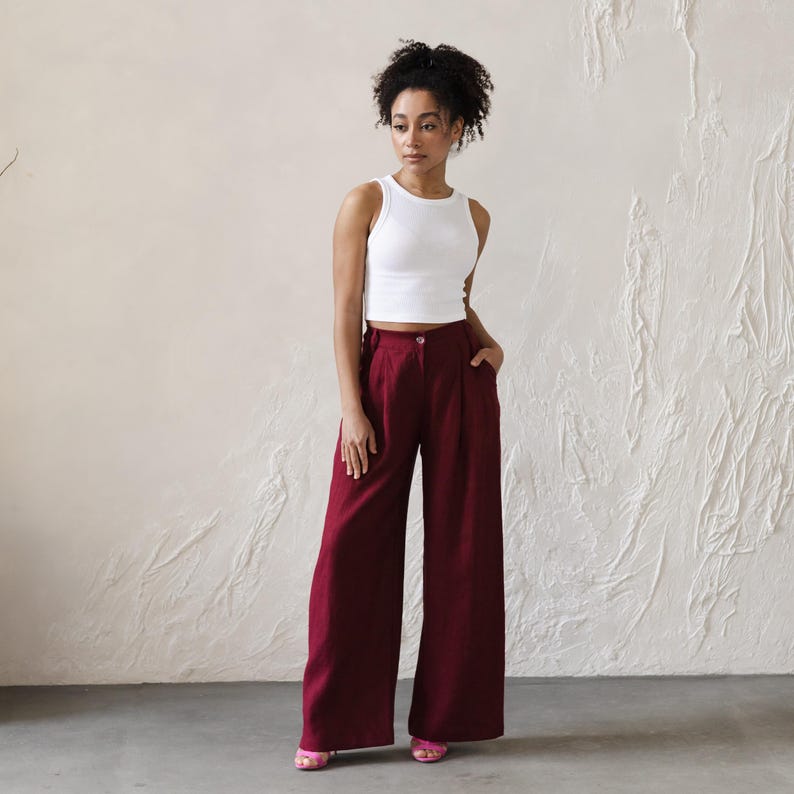 May include: A woman wearing a white tank top and wide-legged burgundy pants. The pants have a high waist and pleats.