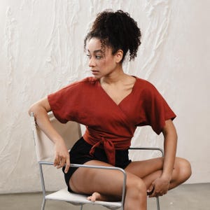 May include: A terracotta-coloured wrap top with short sleeves and a V-neck, tied at the waist. Worn with black shorts. The top is made of a soft, flowing fabric. The model is seated on a modern chair with a white seat and silver frame.