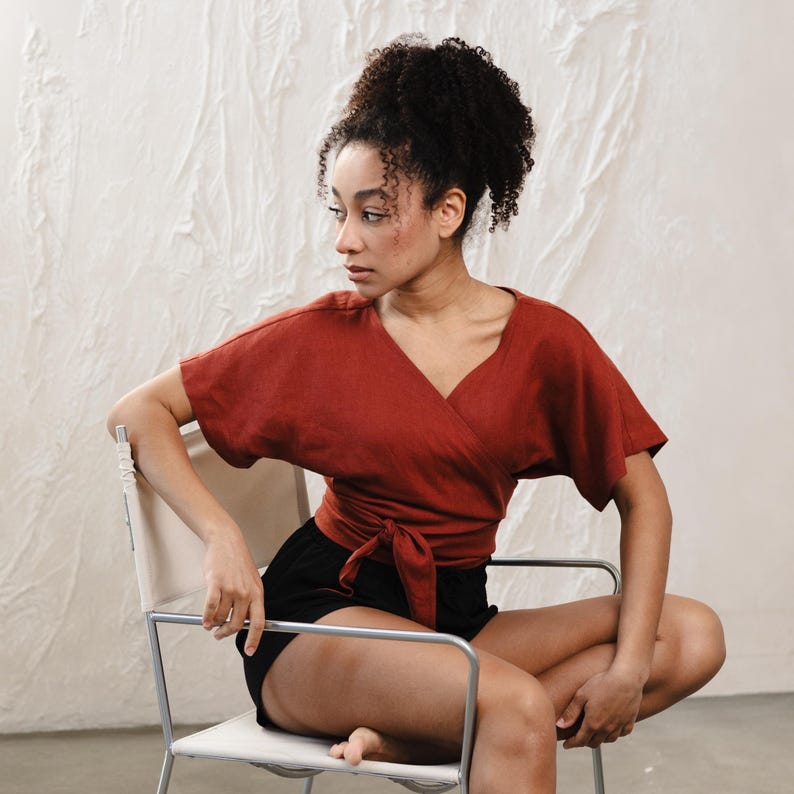 May include: A terracotta-coloured wrap top with short sleeves and a V-neck, tied at the waist. Worn with black shorts. The top is made of a soft, flowing fabric. The model is seated on a modern chair with a white seat and silver frame.