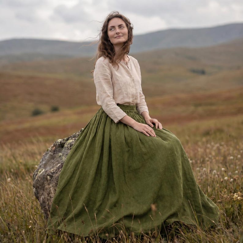 May include: A long-sleeved beige linen shirt with a button-down front and a high-waisted, floor-length olive green linen skirt. The skirt features a full, gathered silhouette that drapes down to the ground, paired with a neutral-toned top.