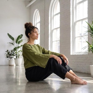 Puede incluir: Una persona con una blusa verde de manga acampanada y pantalones negros sentada en un suelo de hormig&oacute;n. La blusa tiene cuello redondo. El fondo incluye paredes blancas, ventanas arqueadas y plantas en macetas. Los pantalones son holgados.