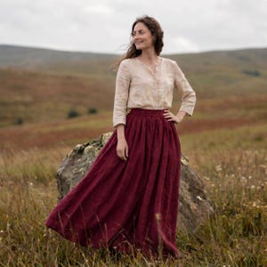 May include: A long, deep burgundy skirt with a gathered elastic waistband and a flowing silhouette, paired with a long-sleeved, light beige button-down shirt featuring a subtle, mottled pattern and a simple round neckline.
