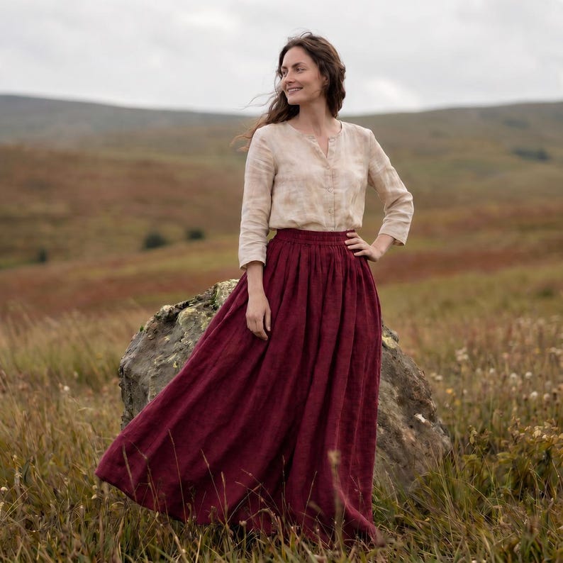 May include: A long, deep burgundy skirt with a gathered elastic waistband and a flowing silhouette, paired with a long-sleeved, light beige button-down shirt featuring a subtle, mottled pattern and a simple round neckline.