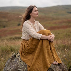 May include: A long-sleeved beige linen blouse with a round neckline and a button-down front, paired with a long, flowing mustard yellow linen skirt. The garments feature a visible natural fabric texture and are worn together as a complete outfit.