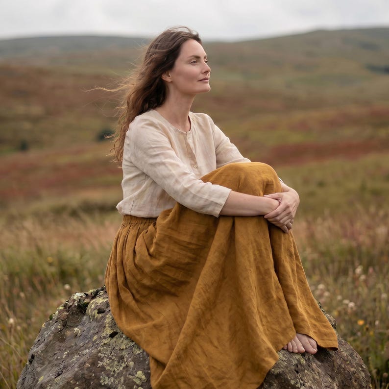 May include: A long-sleeved beige linen blouse with a round neckline and a button-down front, paired with a long, flowing mustard yellow linen skirt. The garments feature a visible natural fabric texture and are worn together as a complete outfit.