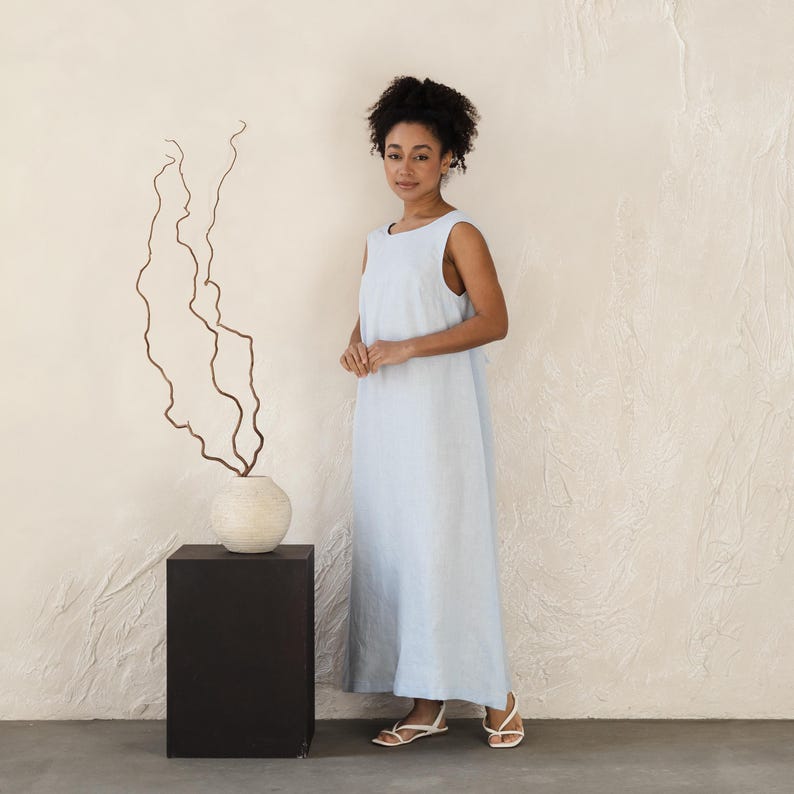 May include: A sleeveless, light blue maxi dress. The dress is worn with white sandals. A woman is standing in front of a textured, off-white wall. A decorative vase with dried branches sits on a black pedestal.