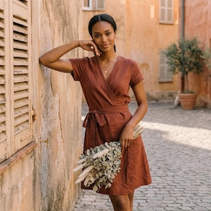 Puede incluir: Un vestido cruzado de color &oacute;xido con mangas cortas y cintura anudada. El vestido est&aacute; hecho de una tela similar al lino y lo sostiene una persona. La persona sostiene un ramo de flores secas.