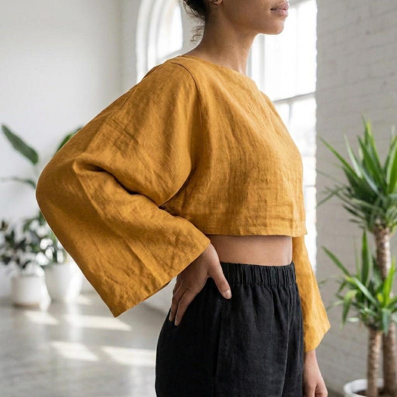 Puede incluir: Top corto de lino amarillo mostaza con mangas largas y anchas. El top se combina con pantalones negros. El conjunto es sencillo y elegante, perfecto para un look casual pero con estilo. La tela parece ligera y transpirable.