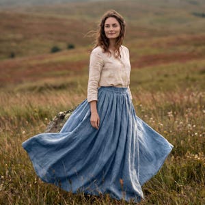 May include: A long-sleeved beige button-down shirt paired with a voluminous, floor-length blue linen skirt. The skirt features a gathered, elasticised waistband and a full, flowing silhouette that extends outwards, showcasing the fabric texture.
