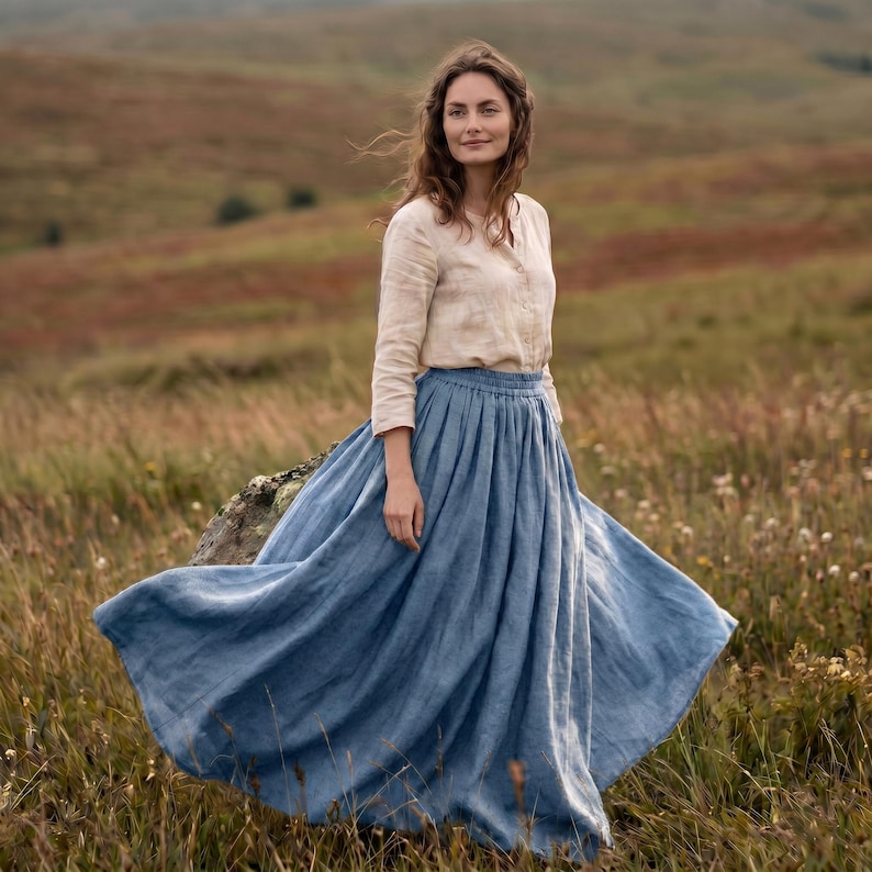 May include: A long-sleeved beige button-down shirt paired with a voluminous, floor-length blue linen skirt. The skirt features a gathered, elasticised waistband and a full, flowing silhouette that extends outwards, showcasing the fabric texture.