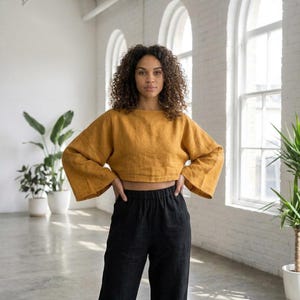 Puede incluir: Un top corto amarillo mostaza de manga larga y ancha, con cuello barco. Se combina con pantalones negros de cintura alta. El conjunto es de tejido suave. El fondo presenta paredes blancas y grandes ventanas.