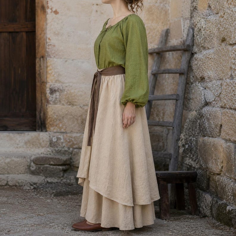 May include: A long-sleeved olive green blouse with a gathered neckline paired with a layered beige maxi skirt. The skirt features an asymmetrical hemline and is cinched at the waist with a dark brown fabric tie belt. The outfit is made of a textured fabric.