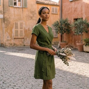 Puede incluir: Una mujer con un vestido cruzado verde oliva de manga corta y cintur&oacute;n a juego, sosteniendo un ramo de flores secas. El vestido tiene cuello en V. El fondo muestra una calle adoquinada y edificios.