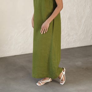 May include: A long, olive green linen dress. The dress is sleeveless and falls to the ankles. White sandals with thin straps and square toes are worn with the dress. The background is a neutral color.